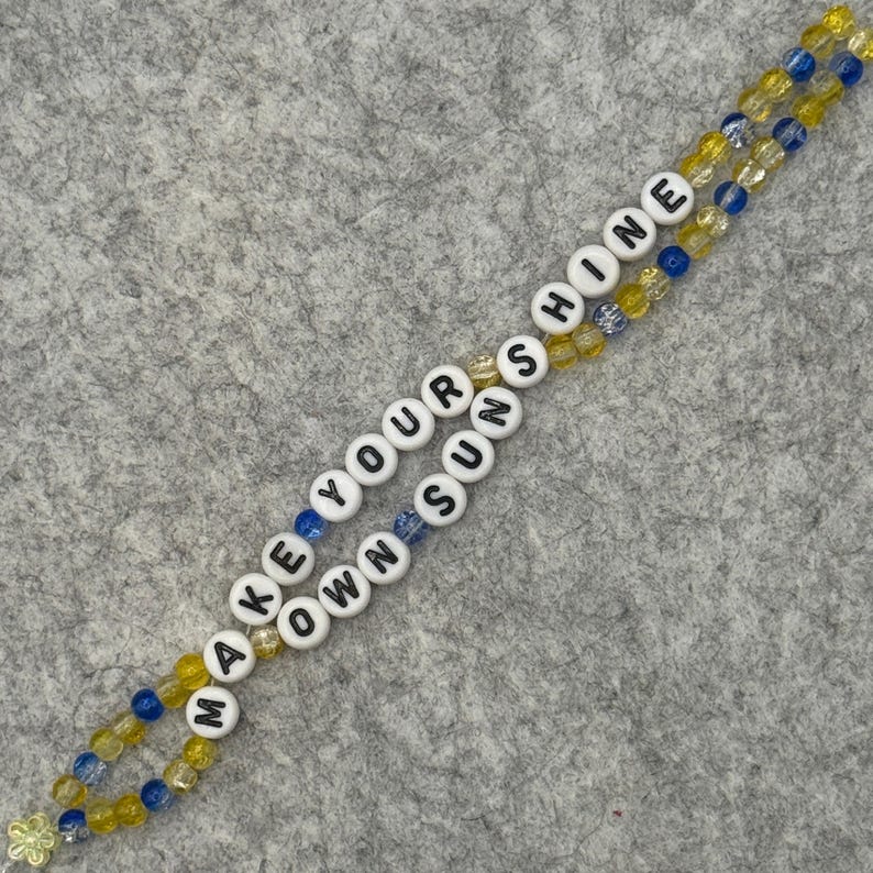 May include: A beaded lanyard with the words "MAKE YOUR OWN SUN SHINE" spelled out in white letter beads. The lanyard features yellow and blue beads, and a small flower charm at the end.