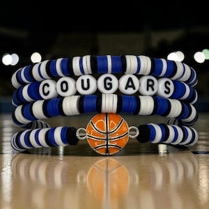 May include: Stack of four bracelets with blue, white, and black beads. One bracelet has the word "COUGARS" spelled out in white letter beads. A basketball charm is attached to one bracelet. The bracelets are on a wooden surface.