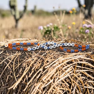 May include: A beaded bracelet featuring alternating orange and blue faceted beads, white round letter beads spelling out I KNEW IT I KNEW YOU, and a single small orange heart bead at one end, arranged in a circular pattern on a textured surface.