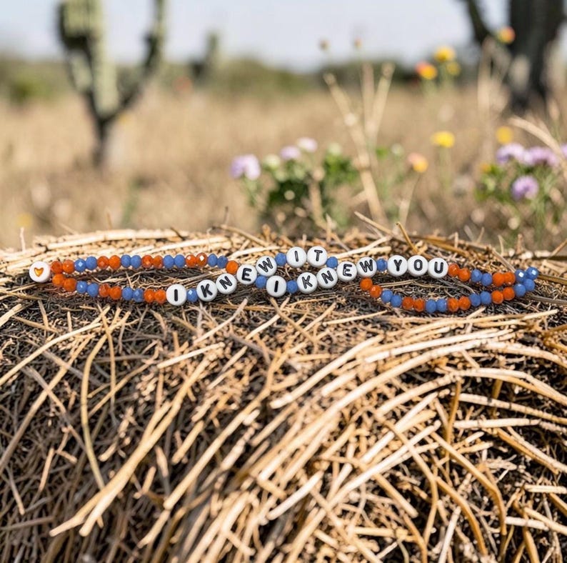 May include: A beaded bracelet featuring alternating orange and blue faceted beads, white round letter beads spelling out I KNEW IT I KNEW YOU, and a single small orange heart bead at one end, arranged in a circular pattern on a textured surface.