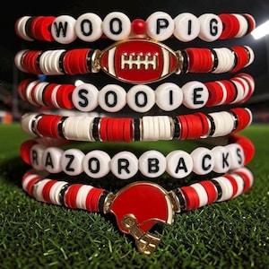May include: A stack of six beaded bracelets featuring red and white disc beads, black spacers, and white letter beads spelling WOO PIG SOOIE RAZORBACKS. The set includes a red and white football charm and a red football helmet charm with gold accents.