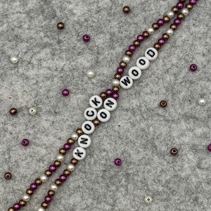 May include: A beaded lanyard with the words "KNOCK WOOD" spelled out in white letter beads. The lanyard features alternating strands of brown, white, and purple beads. Scattered around are additional beads in similar colors.