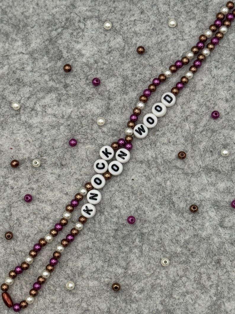 May include: A beaded lanyard with the words "KNOCK WOOD" spelled out in white letter beads. The lanyard features alternating strands of brown, white, and purple beads. Scattered around are additional beads in similar colors.