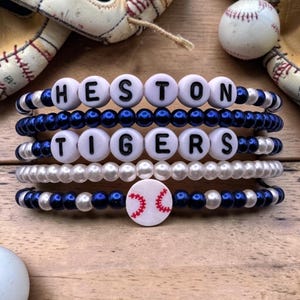 May include: Four beaded bracelets spelling "HESTON TIGERS" with white letter beads. The bracelets feature blue and white beads, and a baseball charm. Baseball gloves and a baseball are in the background, on a wooden surface.