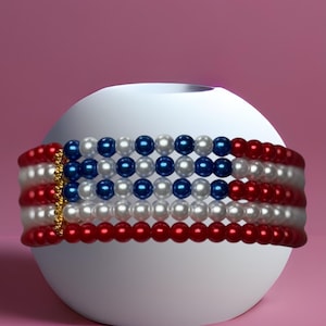 Op de afbeelding: Een kralen choker ketting met een patriottisch ontwerp. De ketting heeft rijen rode, witte en blauwe kralen, die lijken op de Amerikaanse vlag. De ketting is gewikkeld rond een wit bolvormig object tegen een roze achtergrond.