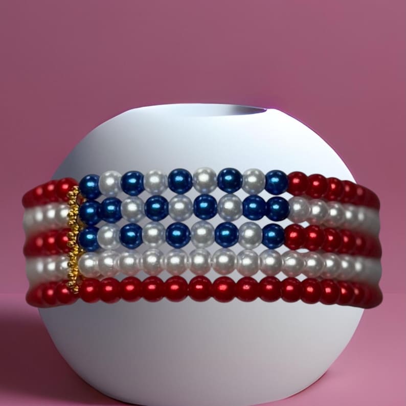 Op de afbeelding: Een kralen choker ketting met een patriottisch ontwerp. De ketting heeft rijen rode, witte en blauwe kralen, die lijken op de Amerikaanse vlag. De ketting is gewikkeld rond een wit bolvormig object tegen een roze achtergrond.