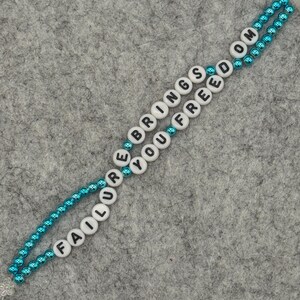 May include: A beaded necklace with the words "FAILURE BRINGS YOU FREEDOM" spelled out in black letters on white beads. The beads are strung on a teal-colored chain, creating a two-strand design. The background is a gray felt surface.