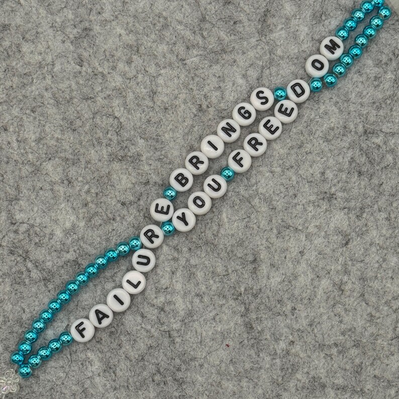 May include: A beaded necklace with the words "FAILURE BRINGS YOU FREEDOM" spelled out in black letters on white beads. The beads are strung on a teal-colored chain, creating a two-strand design. The background is a gray felt surface.