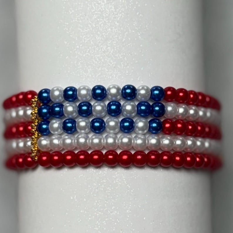 Op de afbeelding: Een kralenarmband met een ontwerp dat doet denken aan de Amerikaanse vlag. De armband heeft afwisselende rijen rode, witte en blauwe kralen, met een goudkleurige sluiting. De armband is ongeveer 1,5 cm breed.