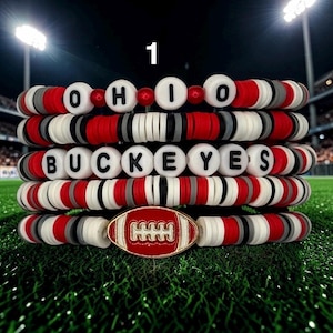 May include: Stack of five bracelets with red, white, gray, and black beads. The top bracelet spells "OHIO" with white letter beads. The third bracelet spells "BUCKEYES". The bottom bracelet features a football charm.