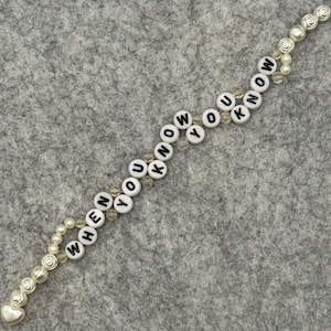 May include: A beaded bracelet with the words "WHEN YOU KNOW" spelled out in black letters on white round beads. The bracelet features pearl and rose-shaped accents, and a heart-shaped charm.
