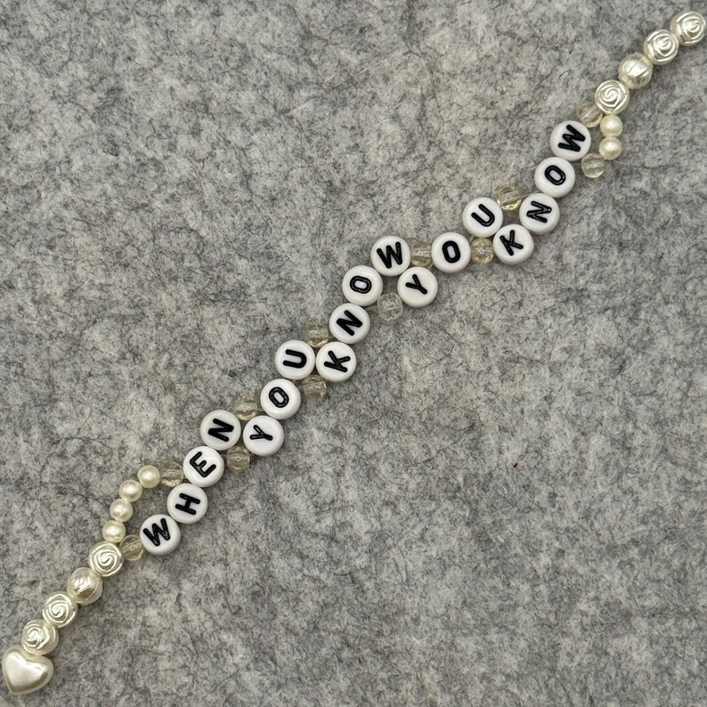 May include: A beaded bracelet with the words "WHEN YOU KNOW" spelled out in black letters on white round beads. The bracelet features pearl and rose-shaped accents, and a heart-shaped charm.