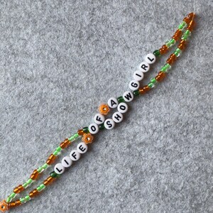 Bracelet made of 2 intertwining strings of 4mm seed shape light green and orange beads with white beads with black letters that say "LIFE OF A SHOWGIRL"