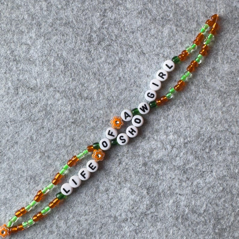 Bracelet made of 2 intertwining strings of 4mm seed shape light green and orange beads with white beads with black letters that say "LIFE OF A SHOWGIRL"
