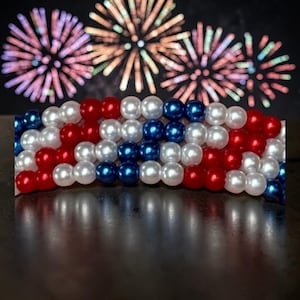 Op de afbeelding: Een patriottische armband gemaakt van rode, witte en blauwe kralen, gerangschikt in een vlagachtig patroon. De kralen zijn rond en glanzend, met een donkere achtergrond met kleurrijk vuurwerk. Een feestelijk accessoire voor patriottische evenementen.