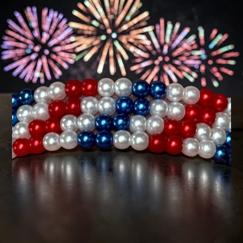 Op de afbeelding: Een patriottische armband gemaakt van rode, witte en blauwe kralen, gerangschikt in een vlagachtig patroon. De kralen zijn rond en glanzend, met een donkere achtergrond met kleurrijk vuurwerk. Een feestelijk accessoire voor patriottische evenementen.