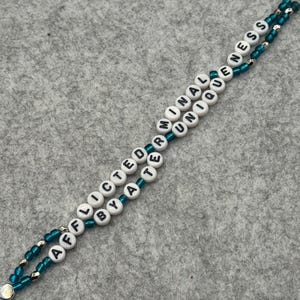 May include: A beaded lanyard with white letter beads spelling "AFFLICTED BY A TERMINAL UNIQUENESS" on a teal cord. The lanyard has silver beads and a silver clasp.