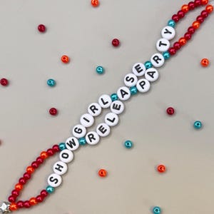 May include: A beaded phone strap with the words "SHOW GIRL RELEASE PARTY" spelled out in white letter beads. The strap features red and orange beads, with teal accents. Scattered around are red, orange, and teal beads.
