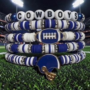 May include: A stack of blue and white beaded bracelets. The top bracelet spells "COWBOYS" in white letter beads. One bracelet has a football charm, another a helmet charm. The bracelets rest on a green surface, resembling a football pitch.