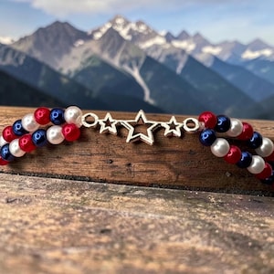 Op de afbeelding: Een patriottische ketting met rode, witte en blauwe kralen, met een zilveren sterhanger. De ketting bestaat uit twee strengen kralen. De achtergrond toont een bergketen. De kleuren doen denken aan een vlag.