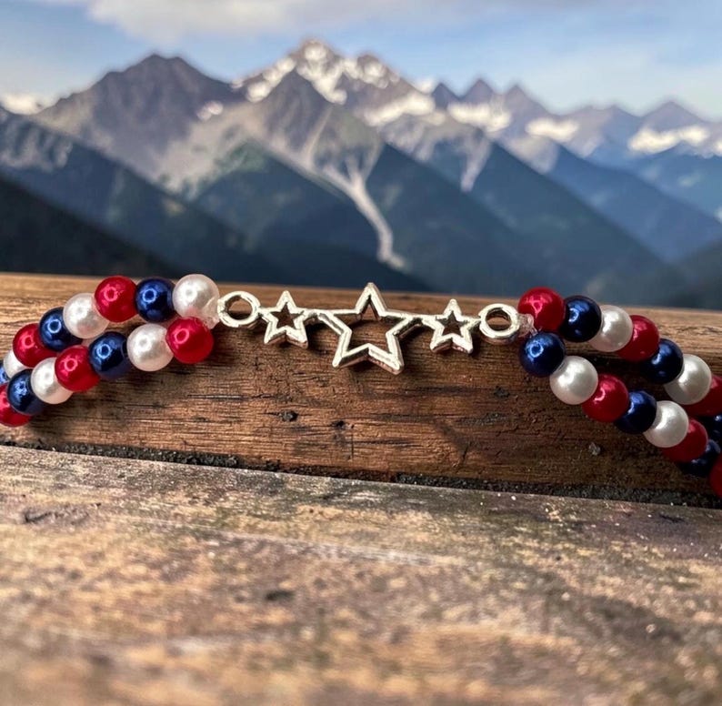 Op de afbeelding: Een patriottische ketting met rode, witte en blauwe kralen, met een zilveren sterhanger. De ketting bestaat uit twee strengen kralen. De achtergrond toont een bergketen. De kleuren doen denken aan een vlag.
