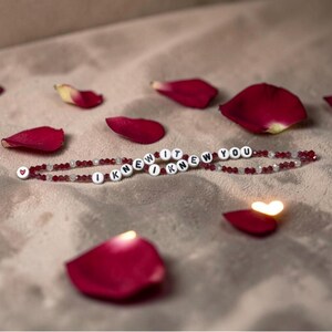 May include: A beaded bracelet featuring alternating dark red and clear faceted beads with white circular letter beads spelling out I KNEW IT I KNEW YOU. The bracelet ends with a small white bead featuring a red heart symbol on one side.