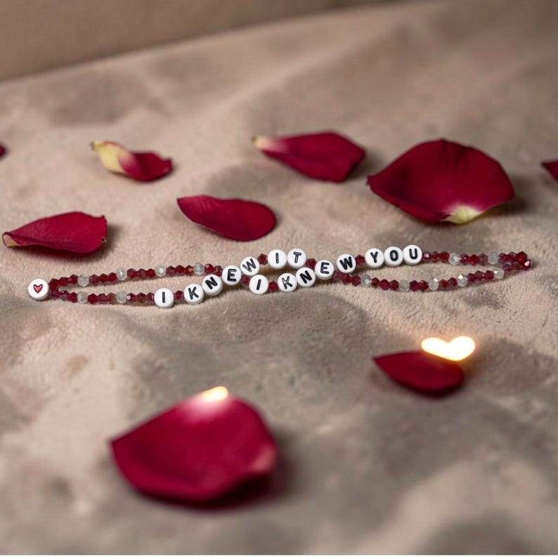 May include: A beaded bracelet featuring alternating dark red and clear faceted beads with white circular letter beads spelling out I KNEW IT I KNEW YOU. The bracelet ends with a small white bead featuring a red heart symbol on one side.