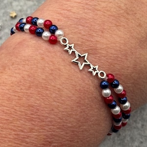 A double-strand bracelet featuring red, white, and blue beads, with a silver-tone 3-star charm. The beads are arranged in a patriotic color scheme. The bracelet is designed to be worn on the wrist.