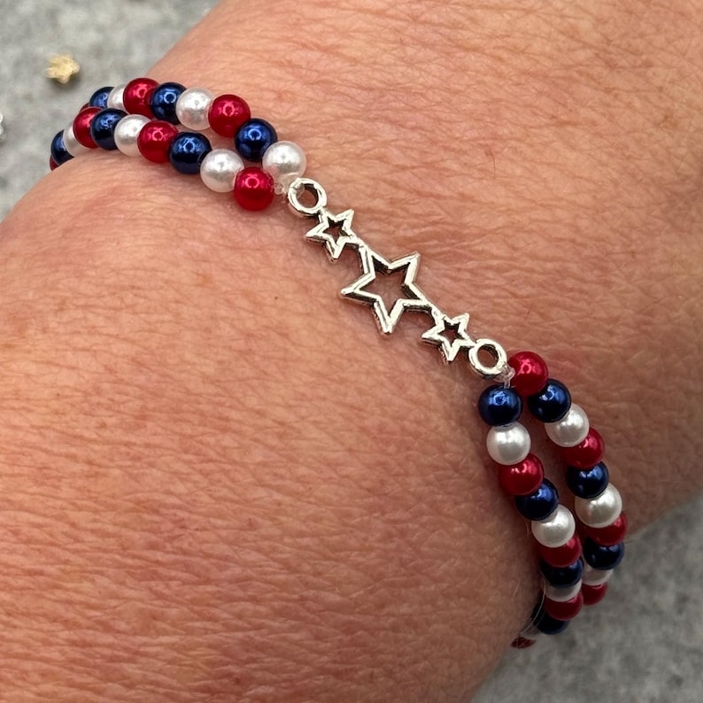 A double-strand bracelet featuring red, white, and blue beads, with a silver-tone 3-star charm. The beads are arranged in a patriotic color scheme. The bracelet is designed to be worn on the wrist.