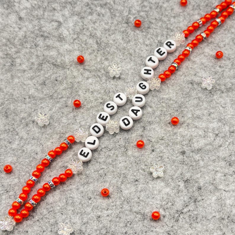 May include: A beaded necklace with the words "ELDEST DAUGHTER" spelled out in white letter beads. The necklace features red beads and clear flower-shaped accents. Additional red beads and flower-shaped accents are scattered around the necklace.