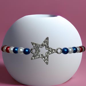 Op de afbeelding: Een armband met een ster-vormige bedel bezet met strass-steentjes. De armband heeft rode, witte en blauwe kralen. Het sieraad wordt getoond op een witte bol tegen een roze achtergrond.