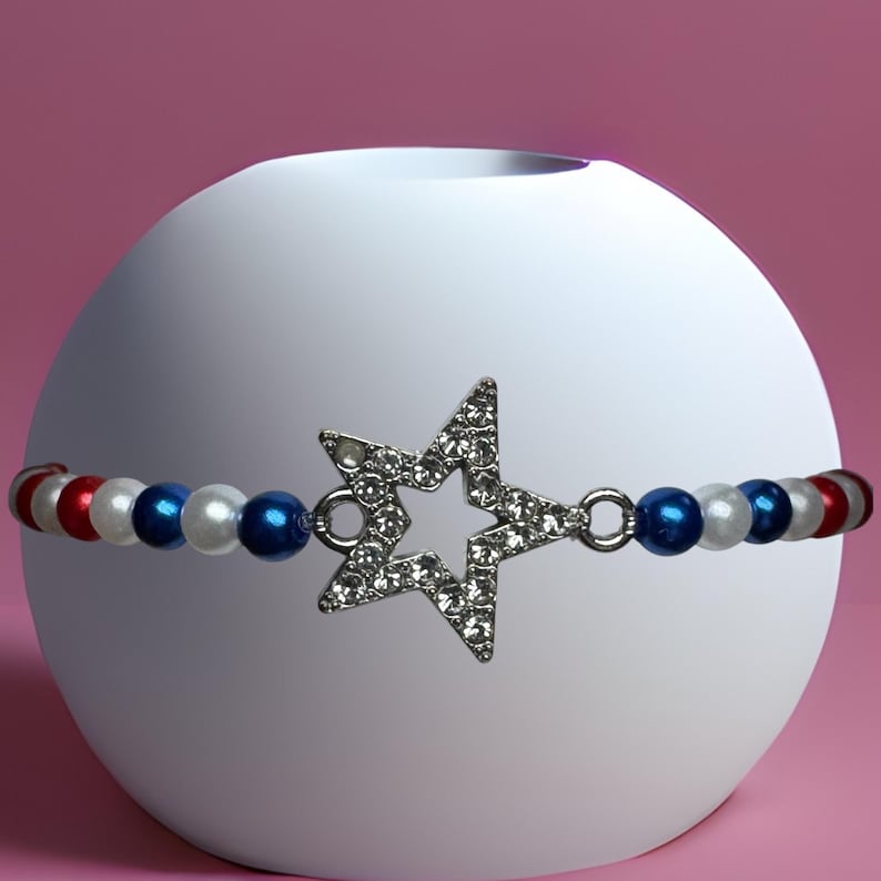 Op de afbeelding: Een armband met een ster-vormige bedel bezet met strass-steentjes. De armband heeft rode, witte en blauwe kralen. Het sieraad wordt getoond op een witte bol tegen een roze achtergrond.