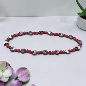 May include: A beaded necklace featuring a repeating pattern of small, faceted red beads interspersed with metallic, chevron-shaped gray beads. The necklace is arranged in an oval shape, showcasing a consistent sequence of red and gray components.