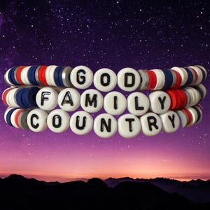 Op de afbeelding: Drie gestapelde armbanden met de woorden "GOD FAMILY COUNTRY" in zwarte letters op witte kralen. De armbanden hebben rode, witte en blauwe accentkralen, tegen een paarse en zwarte achtergrond.