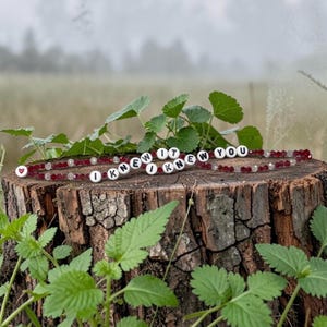 May include: A beaded bracelet featuring deep red and clear faceted beads, interspersed with white circular letter beads that spell out the phrase I KNEW IT I KNEW YOU, finished with a small white bead containing a red heart symbol.