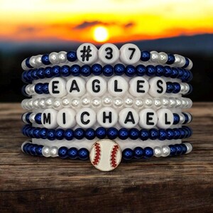 May include: Stack of beaded bracelets with white, navy blue, and silver beads. The bracelets spell out "#37 EAGLES MICHAEL" with white letter beads. A baseball charm is at the base. The background shows a sunset.