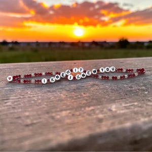May include: Two beaded bracelets featuring small, faceted dark red and clear crystals. Each bracelet includes white circular beads with black lettering that spells out the phrase I KNEW IT I KNEW YOU, with a small red heart bead at one end.