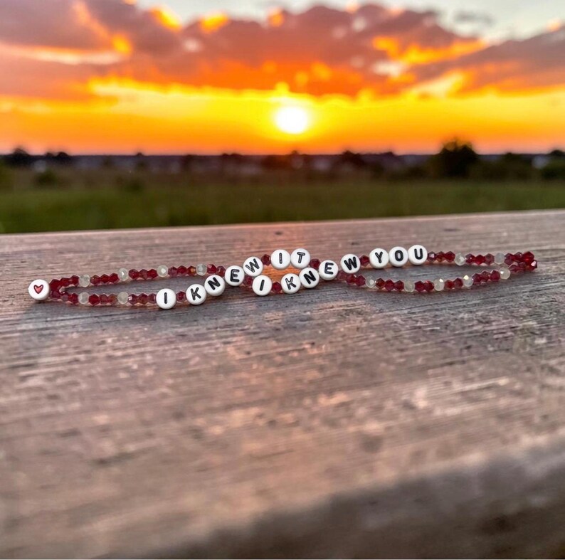 May include: Two beaded bracelets featuring small, faceted dark red and clear crystals. Each bracelet includes white circular beads with black lettering that spells out the phrase I KNEW IT I KNEW YOU, with a small red heart bead at one end.