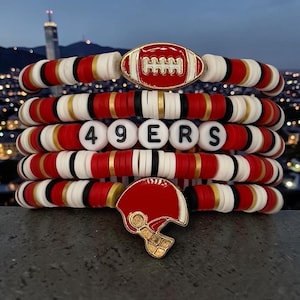 May include: A stack of beaded bracelets in red, white, black, and gold. The bracelets feature a football charm and a helmet charm. One bracelet spells out "49ERS" with white letter beads. The background is a blurred cityscape.