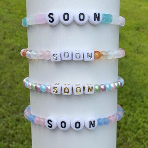 Peut inclure: Quatre bracelets de perles avec le mot "SOON" épelé en perles de lettres blanches. Les bracelets présentent des perles de couleurs pastel, notamment roses, bleues et irisées. Les bracelets sont exposés sur un objet cylindrique blanc.