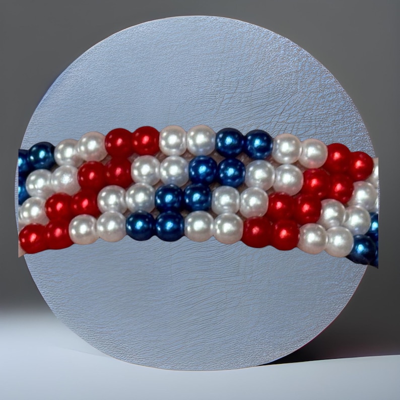 Op de afbeelding: Een rond, lichtgrijs object met een kralenontwerp in rood, wit en blauw, dat op een vlag lijkt. De kralen zijn in horizontale rijen gerangschikt, waardoor een patriottisch patroon ontstaat. De achtergrond is een grijstint.