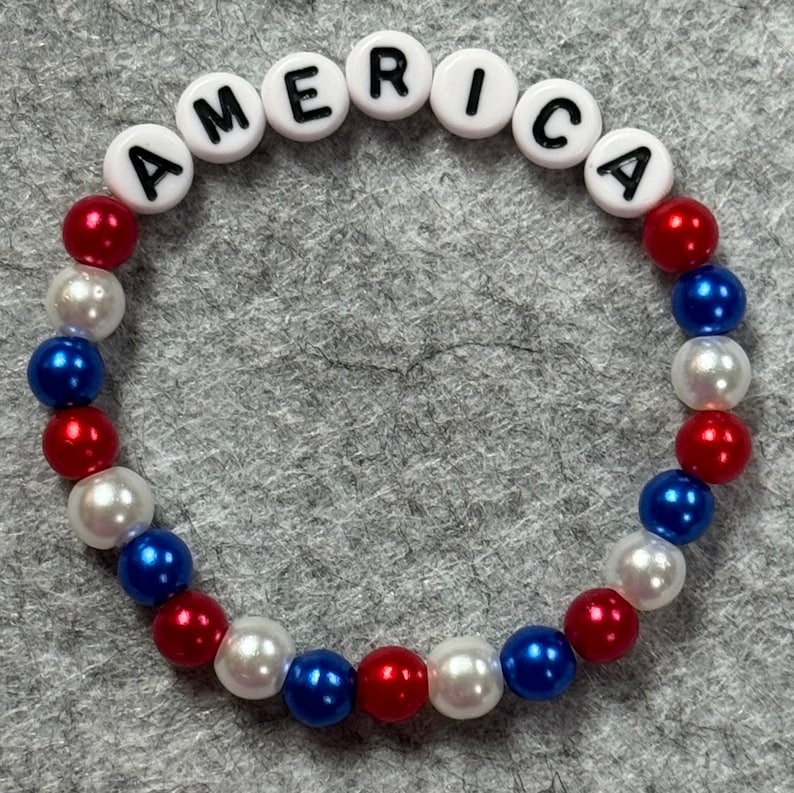 A patriotic bracelet with the word "AMERICA" spelled out in white letter beads. The bracelet features alternating red, white, and blue beads, creating a festive and colorful accessory.