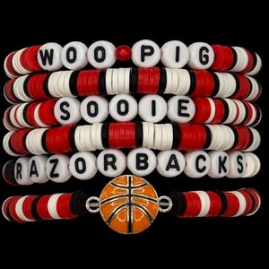 May include: A collection of beaded bracelets in red, white, and black. The bracelets spell out "WOO PIG SOOIE RAZORBACKS" with white letter beads. A basketball charm is featured on one bracelet, with a black background.
