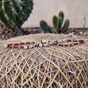 May include: A beaded bracelet featuring dark red faceted crystals and white spherical beads with black lettering that spells out the phrase I KNEW IT I KNEW YOU, finished with a small red heart symbol at one end of the beaded strand.