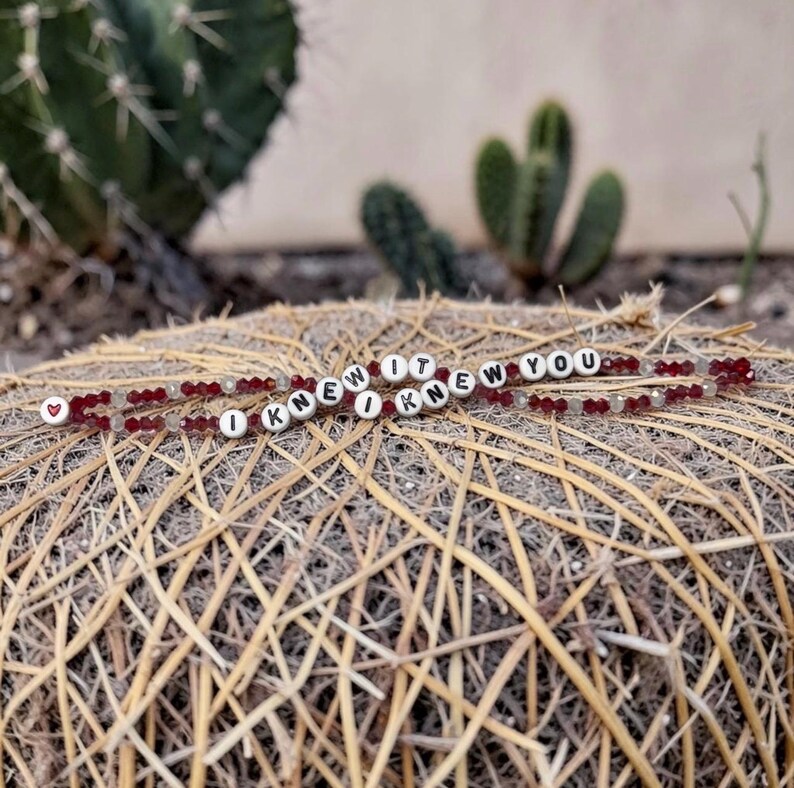 May include: A beaded bracelet featuring dark red faceted crystals and white spherical beads with black lettering that spells out the phrase I KNEW IT I KNEW YOU, finished with a small red heart symbol at one end of the beaded strand.