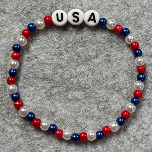A Patriotic beaded bracelet featuring red, white, and blue beads, with the letters "USA" spelled out in white letter beads. The bracelet is designed with alternating colors and is made with elastic string.