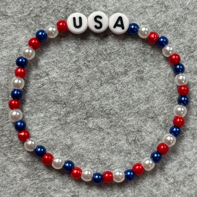 A Patriotic beaded bracelet featuring red, white, and blue beads, with the letters "USA" spelled out in white letter beads. The bracelet is designed with alternating colors and is made with elastic string.