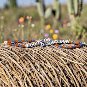 May include: A beaded bracelet featuring alternating orange and blue faceted beads, interspersed with white round beads displaying the black text I KNEW IT I KNEW YOU, and a single orange heart-shaped bead at the end of the strand.