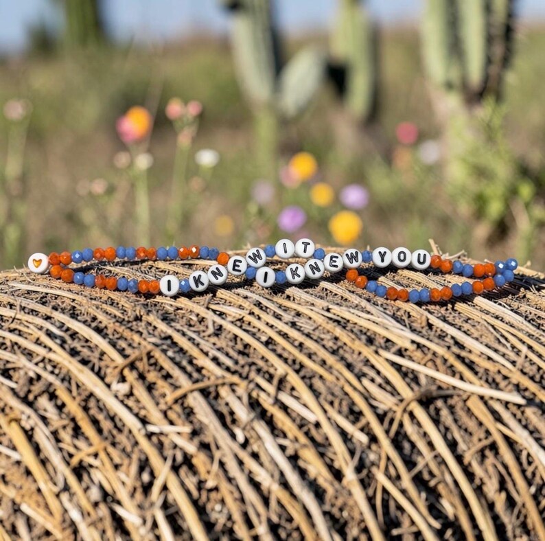 May include: A beaded bracelet featuring alternating orange and blue faceted beads, interspersed with white round beads displaying the black text I KNEW IT I KNEW YOU, and a single orange heart-shaped bead at the end of the strand.