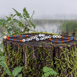 May include: A beaded bracelet featuring alternating orange and blue faceted beads, interspersed with white round beads displaying the black text I KNEW IT I KNEW YOU, and a single small orange heart bead at the end of the beaded strand.